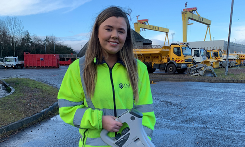 Civil Engineering Apprentice photograoherd wearing a high vis jacket and standing in a yard with vehicles in the background.